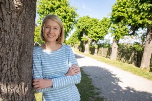 Christine Haberlander in der Natur neben einem Baum mit verschränkten Armen