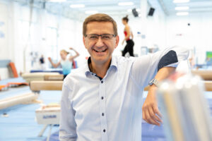 Portrait von Markus Achleitner in einer Sporthalle mit Leichtathletikerinnen im Hintergrund